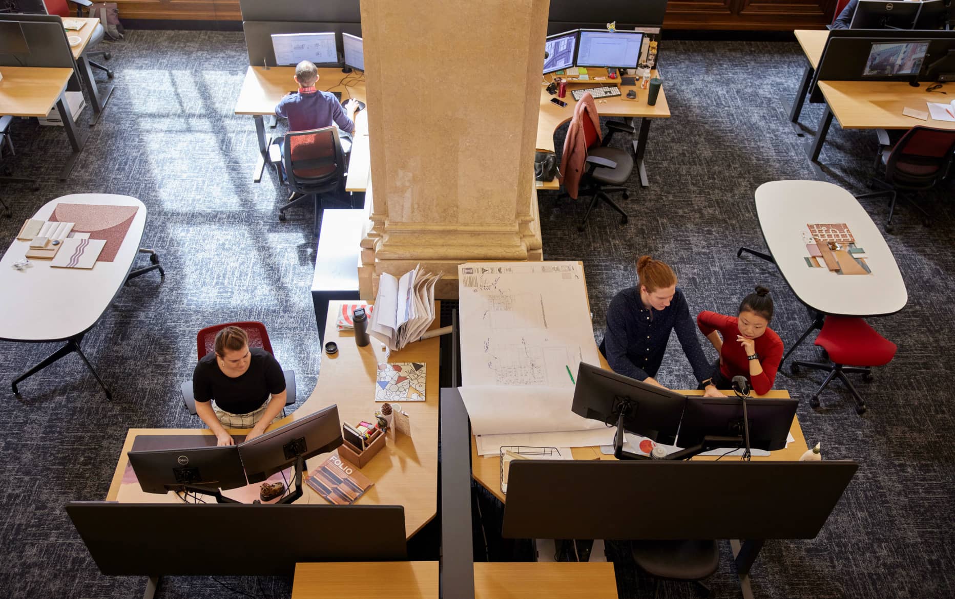 A man and a woman are working at a standing desk, looking at the same monitors, while others are seated at their own desks working.