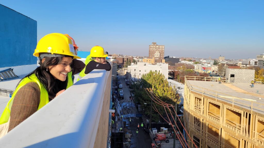 People wearing safety helmets and vests are looking down from the rooftop. A woman at the front is smiling.