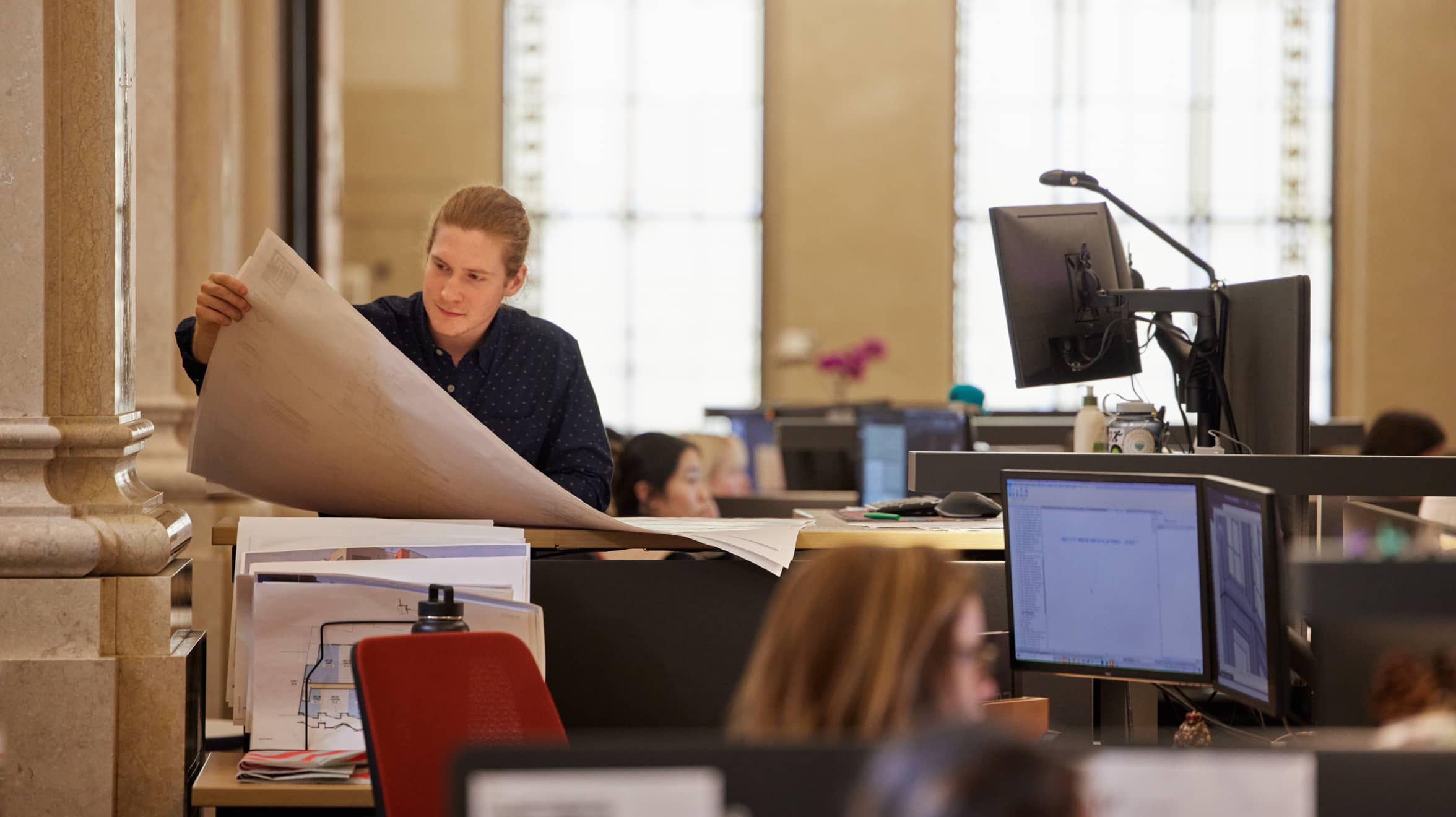 A man is standing at a standing desk, looking at blueprints, while others are seated and looking at an own screen.