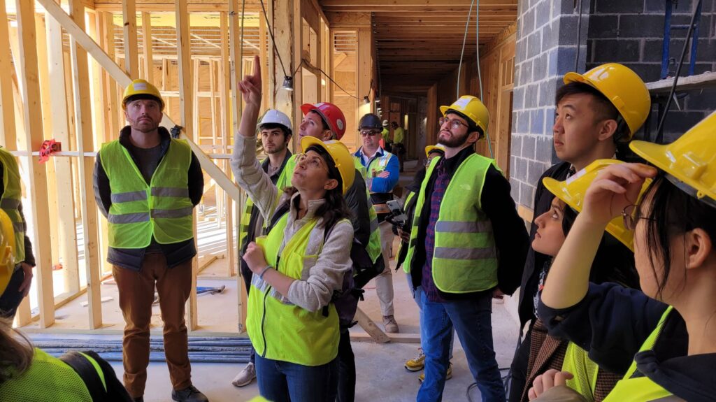 At a construction site, a woman in the middle pointing at something above and people wearing safety helmets are looking upward