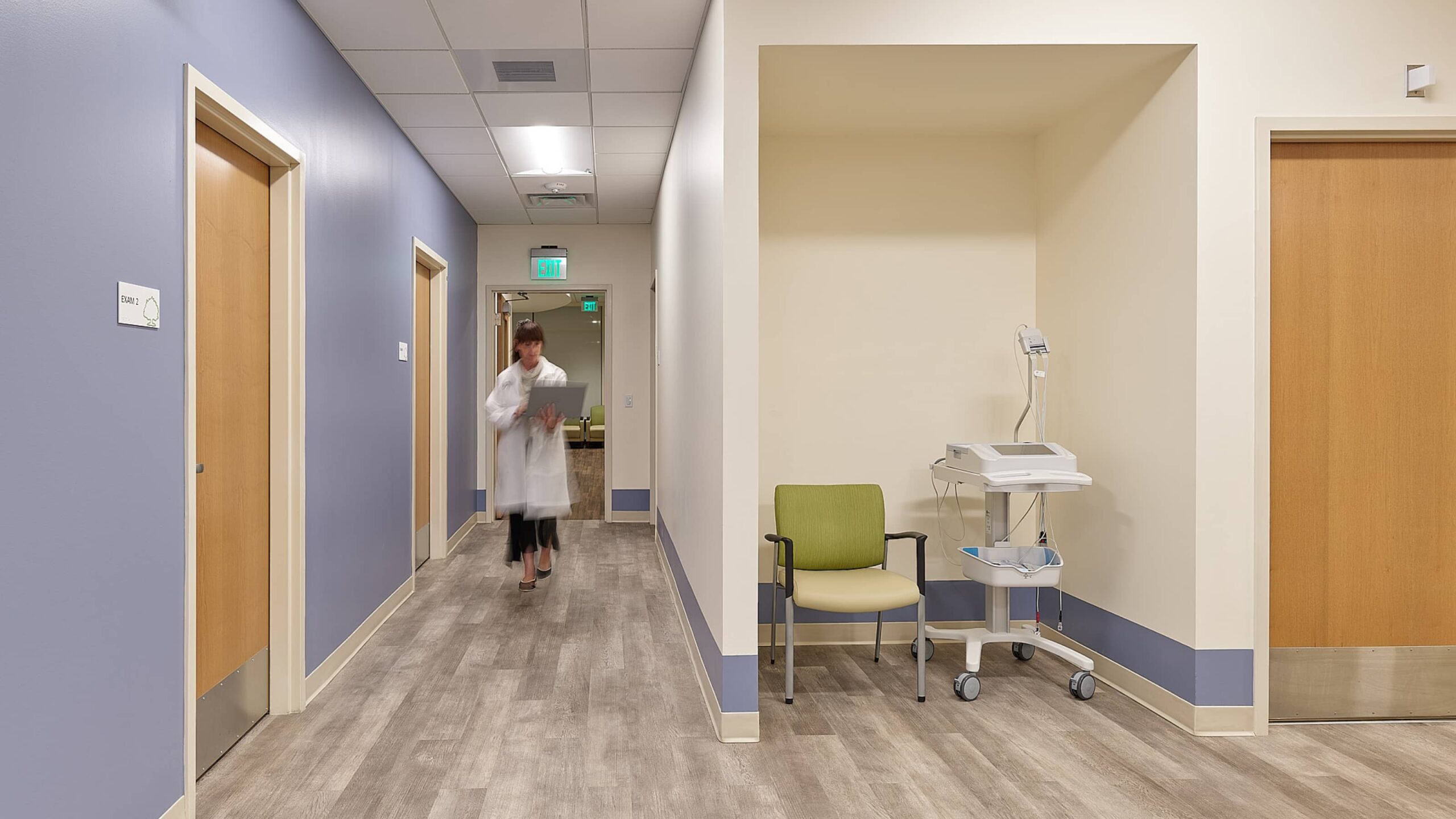 A woman in a doctor's gown is walking through the aisle, holding a laptop. Medical equipment and a chair are situated in an alcove