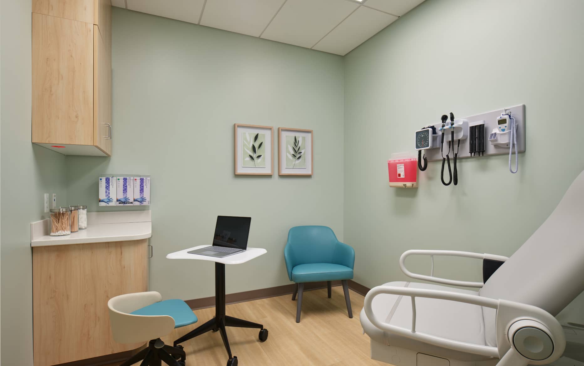 A clean, modern medical examination room with a laptop on a movable desk, a teal chair, an examination bed, wall-mounted medical tools, and minimal decor.