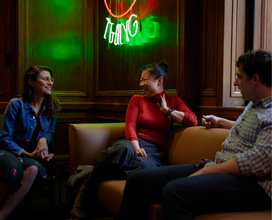 Three people are sitting on a sofa, talking to each other and smiling. A neon sign is hanging on the wall