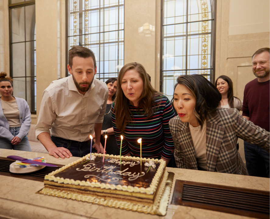 Three people are blowing out candles on the birthday cake, while others are smiling behind them.