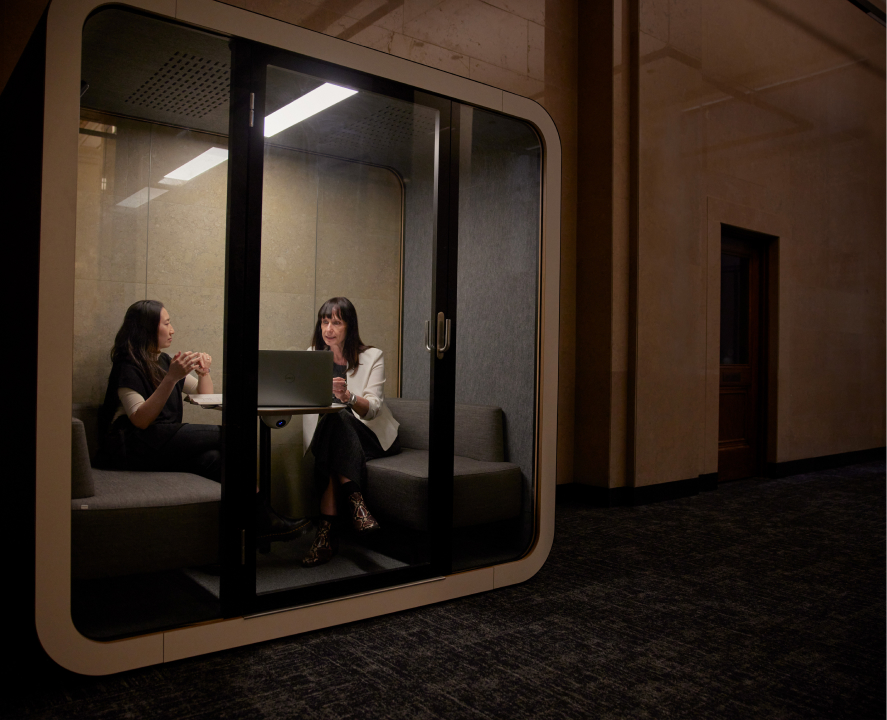Two women are discussing in a meeting cube with a laptop on the desk