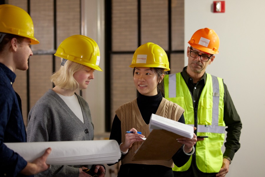 A woman holding documents is talking to other people, all of whom are wearing safety helmets.