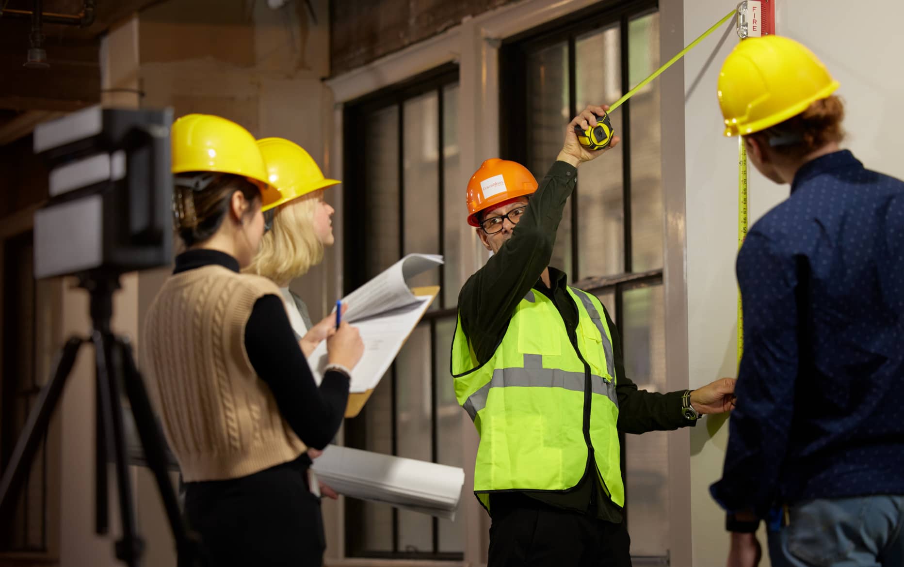 A man in an orange safety helmet is measuring a wall with a tape measure, while three others in yellow safety helmets are observing him and taking notes