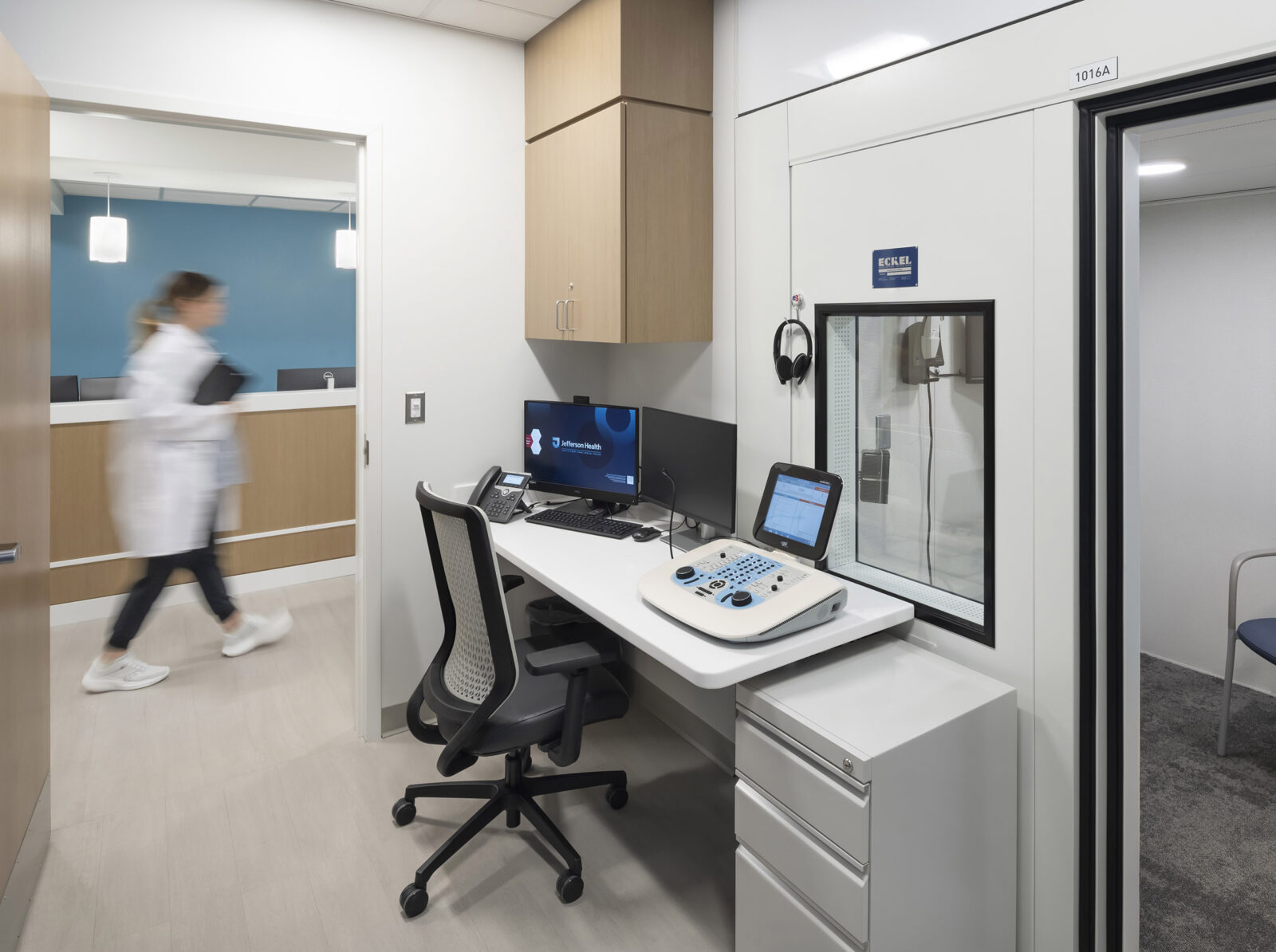 photo of the clinic's audiology suite. a desk and audio equipment sit outside a sound isolated chamber, while a doctor walks by in the hallway